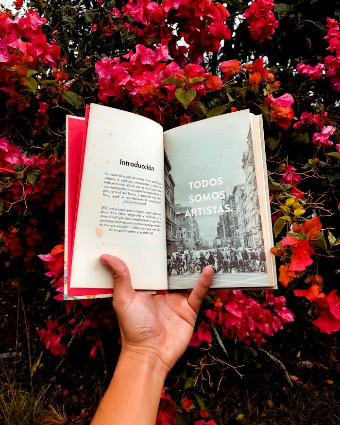 A hand holds an open book with text against a backdrop of vivid pink flowers.